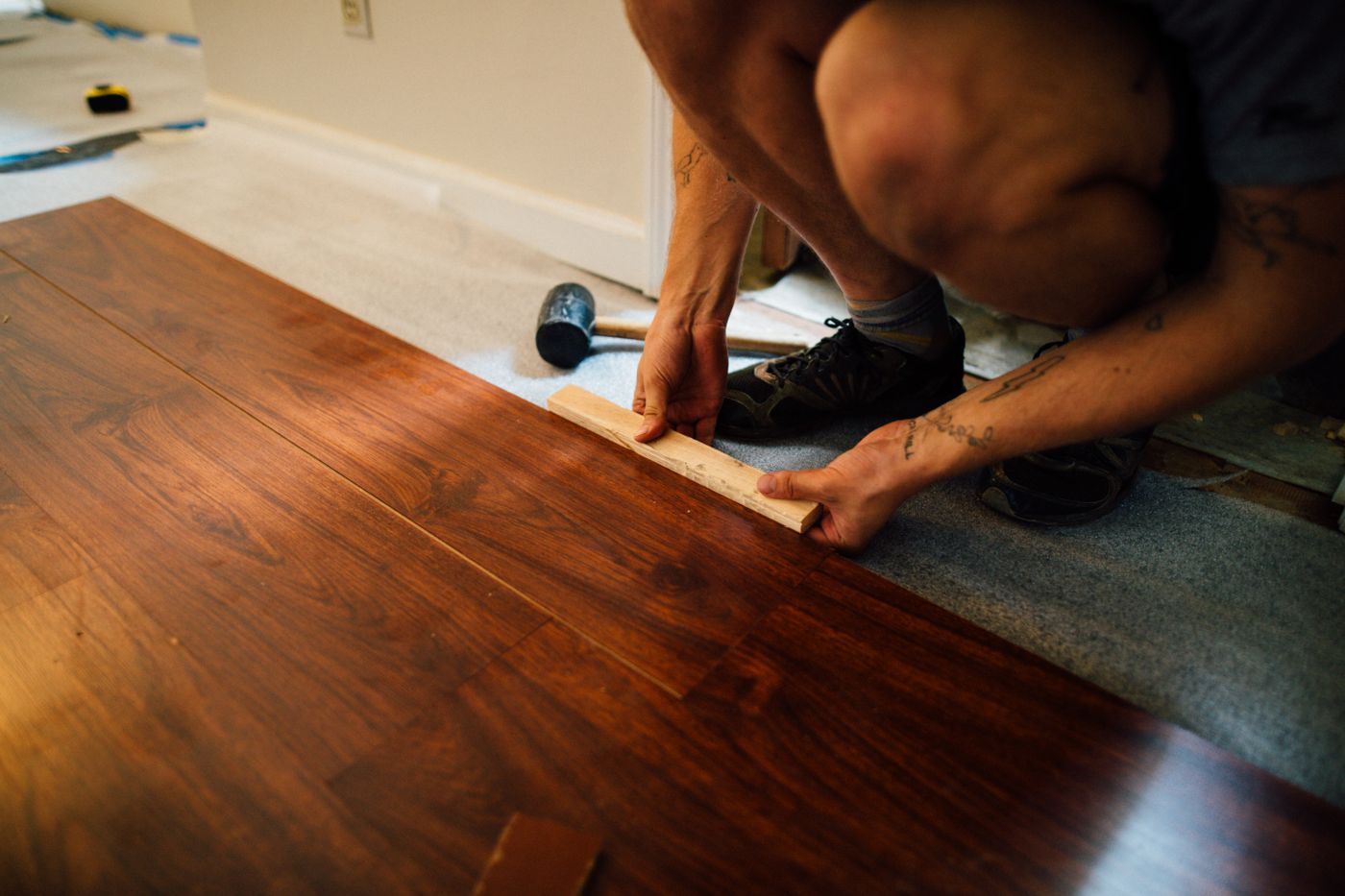 Person fitting a wood-look flooring plank at the edge of a room, with carpet still in place beside it.